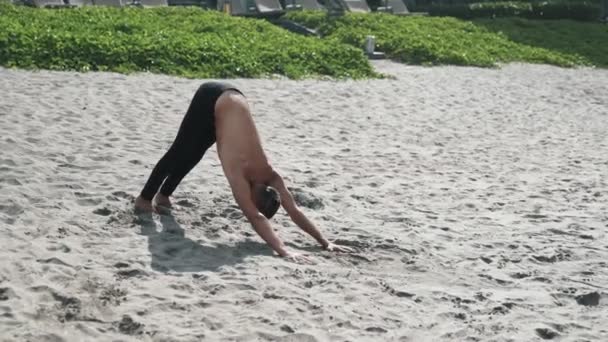 Hombre de pie en pose de yoga y haciendo ejercicios de estiramiento en la playa de arena, steadicam shot — Vídeos de Stock