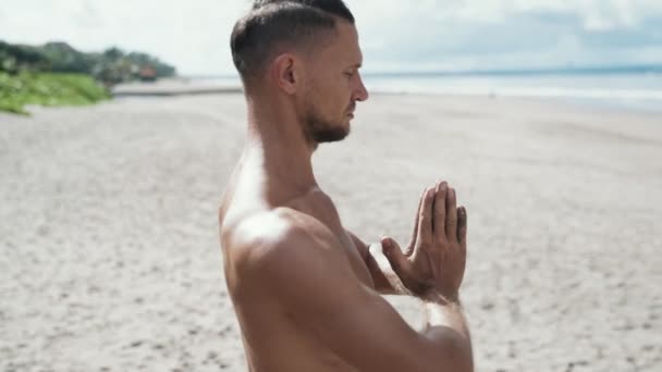 Close-up slow motion steadicam shot, athletic man doing yoga on beach with his hands together and eyes closed — Stock Video