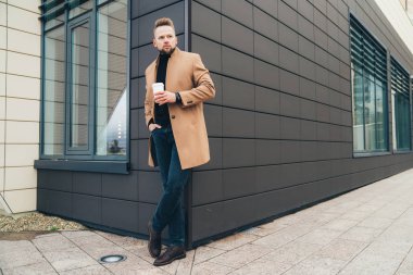 Çekici bir adam trendy kahverengi ceket ve iş merkezine yakın poz elinde kahve