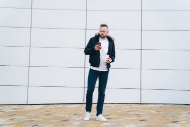 Çekici genç casual elbise kahve ile adam ve ofis binası arka plan ayakta onun elinde telefon.