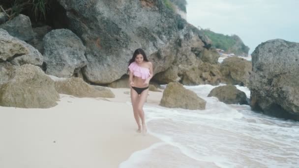 Slow-motion, sexy meisje in een badmode loopt langs het strand met wit zand en grote golven, prachtige rotsen en stenen op de achtergrond. Steadicam schot. — Stockvideo