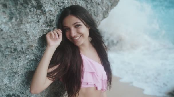 Close-up portrait of smiling beautiful girl, posing near the rock on the beach with big waves. Slow-motion — Stock Video