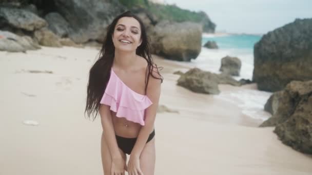 Slow-motion, glimlachend meisje in badmode wandelingen aan zandstrand met grote stenen en golven op de achtergrond, steadicam schot — Stockvideo