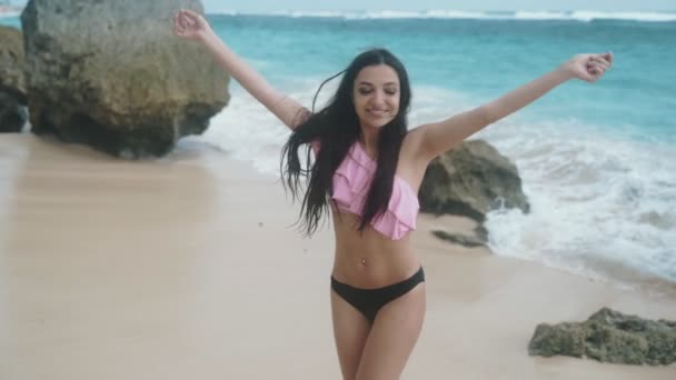Медленное движение счастливая девушка в купальнике вращается вокруг себя с протянутыми руками на фоне волн на пляже, устойчивый снимок — стоковое видео