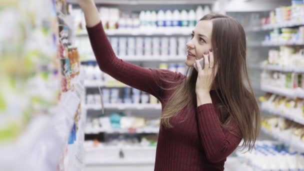 Jolie fille souriante, parlant au téléphone et faisant du shopping dans un supermarché — Video