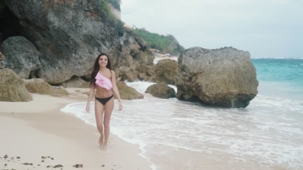 Lassú mozgás szép karcsú lány a fürdőruhát, séta a strand hátteret, az óceán hullámai és a nagy sziklák — Stock videók