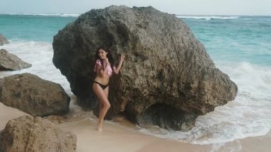 Ağır çekim güzel genç kız mayo ayakta yakınındaki plaj ve arka plan okyanus dalgaları, steadicam vurdu, büyük taş