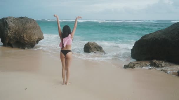 Slow motion achterkant veiw slim meisje in badmode met wijd open armen staat op strand, kijkt naar de oceaan golven, steadicam schot. — Stockvideo