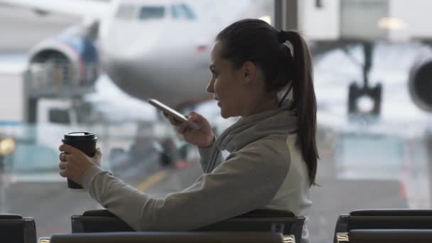 Девушка с кофе на вынос в руке записывает голосовое сообщение по телефону в аэропорту — стоковое видео