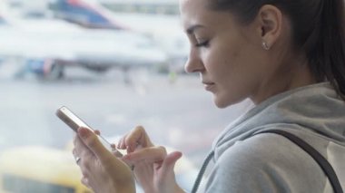 Akıllı telefon, mesaj mesaj penceresinde, uçağın Havaalanı planda kullanan yan görünümü çekici kız yakın çekim. Uçuş için bekleyen kadın. Kavram seyahat, seyahat, Tur, gezi