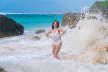 Şehvetli güzel kız gülümseyerek ve kayalar ve dalgalar arka plan ile sahilde poz mayo