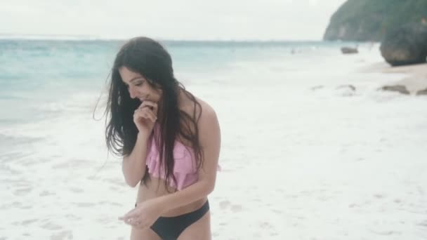 Повільні рухи, чуттєва красива дівчина в купальнику посміхається і позує на пляжі з камінням і хвилями фону, стрімкий постріл — стокове відео