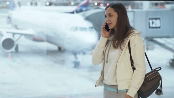 Meisje in sportkleding en met rugzak permanent bij raam in luchthaven en praten over de telefoon achtergrond van vliegtuig — Stockvideo