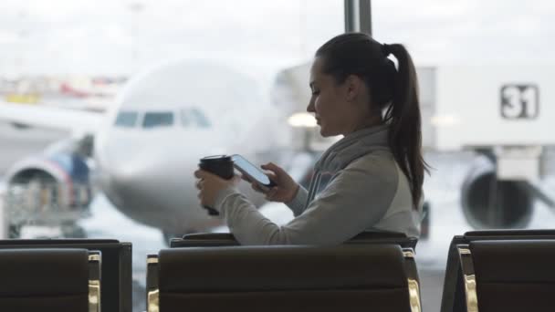Schönes Mädchen trinkt Kaffee zum Mitnehmen und benutzt Telefon, um Nachricht im Flughafen-Wartezimmer zu senden — Stockvideo