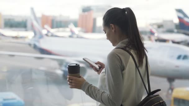 Kant weergave meisje maakt gebruik van telefoon en drankjes afhaalmaaltijden koffie in luchthaven achtergrond van vliegtuig in venster — Stockvideo