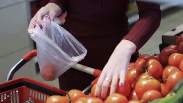 Close up womans hands puts tomatos in plastic bag from counter with fresh vegetables — Stockvideo