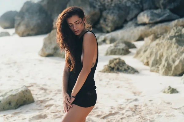 Retrato de modelo morena sexy en suéter de punto posando en la playa de arena, piedras grandes y roca sobre fondo . —  Fotos de Stock