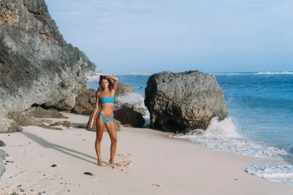 Slanke gebruinde meisje in blauwe zwembroek met opgeheven armen permanent aan zandstrand met grote stenen — Stockfoto