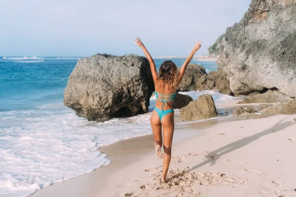 Baksidan syn på smal flicka i baddräkt med upphöjda armar upp står på sandstrand med klippor — Stockfoto