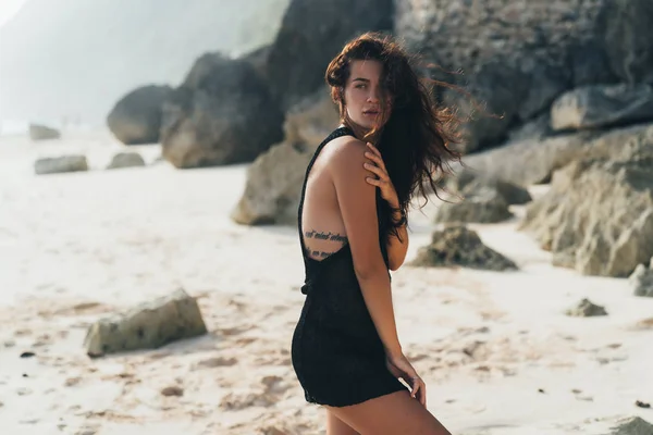 Retrato de modelo morena sexy en suéter de punto posando en la playa de arena, piedras grandes y roca sobre fondo . —  Fotos de Stock