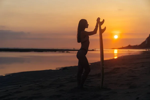 Silueta de chica delgada con tabla de surf en las manos en la playa en el fondo de la hermosa puesta de sol — Foto de Stock