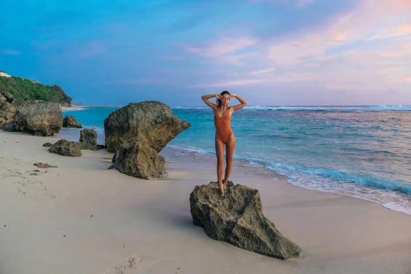 Chica atractiva europea en traje de baño marrón de pie en la playa rocosa durante la puesta del sol — Foto de Stock