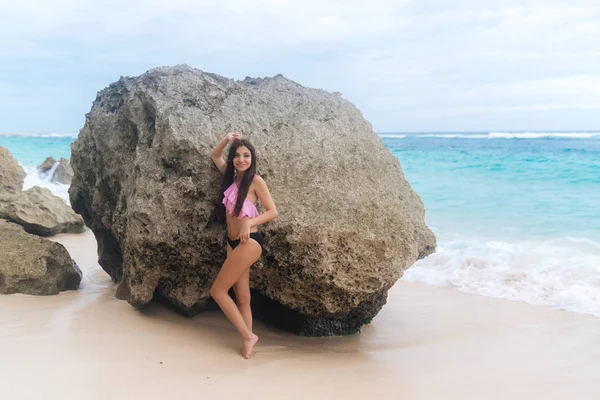 Σέξι μαυρισμένο κορίτσι με μαγιό στέκεται στο φόντο παραλία, μεγάλες πέτρες και ωκεανό — Φωτογραφία Αρχείου