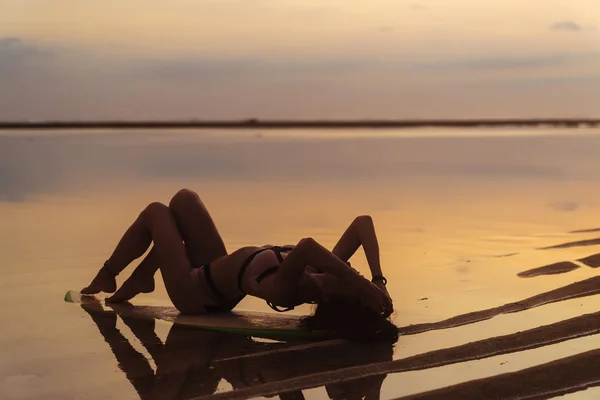 Silhouet sexy meisje in zwembroek liegen en poseren op de surfplank op strand tijdens zonsondergang — Stockfoto