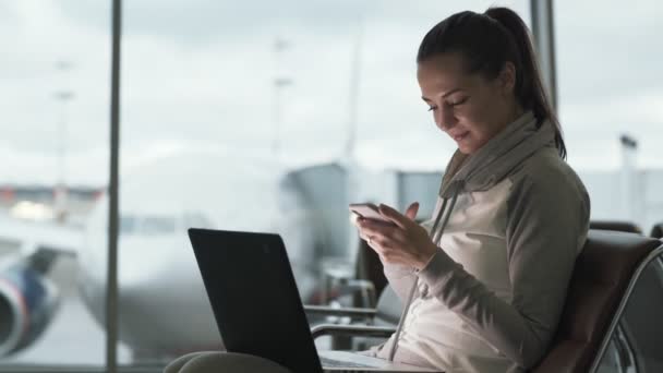Widok z boku z ładną dziewczyną wykorzystuje telefon i laptop do pracy podczas oczekiwania na pokład w odlotów na lotnisku — Wideo stockowe