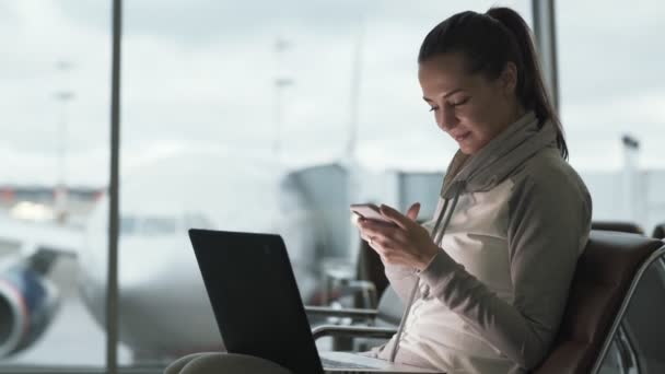 Side view of pretty girl uses phone and laptop to work at airport while waiting boarding at departure lounge — Stock Video