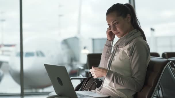 Дівчина мандрівник фрілансер одягає навушники і використовує ноутбук для роботи в залі аеропорту — стокове відео
