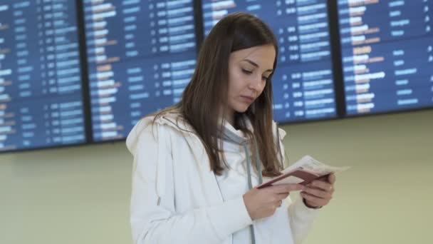 Красивая девушка с билетом и паспортом в руках проверяет время вылета с помощью информационного табло в аэропорту — стоковое видео