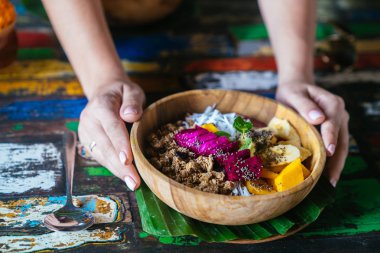 Kadının eller güler yüzlü kase tutarak yapılan mango, muz, granola, rendelenmiş hindistan cevizi, dragon meyve ve renkli arka plan üzerinde nane