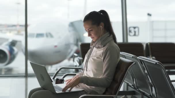 Freiberuflerin mit Kopfhörer in den Ohren, Laptop für Fernarbeit und Warten auf ihren Flug am internationalen Flughafen — Stockvideo