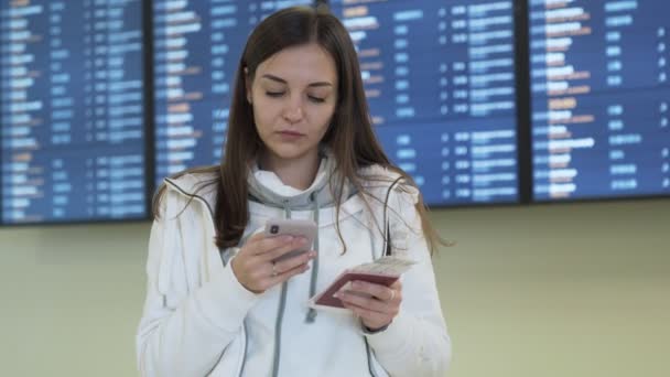 Hermosa chica con pasaporte y billete en sus manos utiliza teléfono y comprueba el horario de salida en el tablero de información en el aeropuerto — Vídeos de Stock