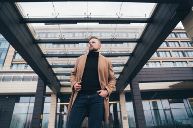 İş merkezine yakın poz trendy kahverengi kabanlı çekici bir adam. Sakallı yakışıklı Avrupa adam uzakta görünüyor. Kavramı yaşam tarzı, moda erkek tarzı