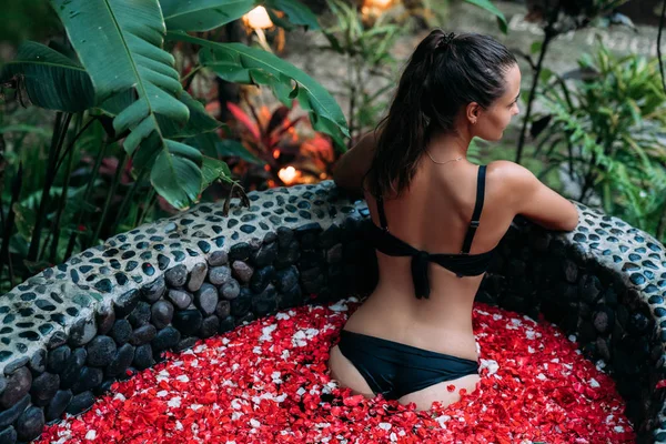Vista trasera de la joven mujer relajándose en el baño con pétalos de rosa y flores en los trópicos — Foto de Stock