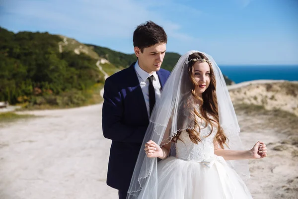 Elegante sposo in abito sta dietro la sposa in bellissimo vestito bianco in posa su roccia con mare e montagne sfondo — Foto Stock