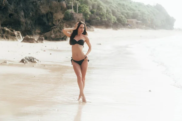 Красивая девушка в черном купальнике гуляет по белому песчаному пляжу возле океана . — стоковое фото