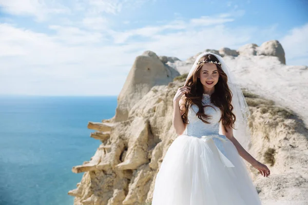 Szép boldog menyasszony, esküvői ruha, és pózol a rock háttér óceán fátyol — Stock Fotó
