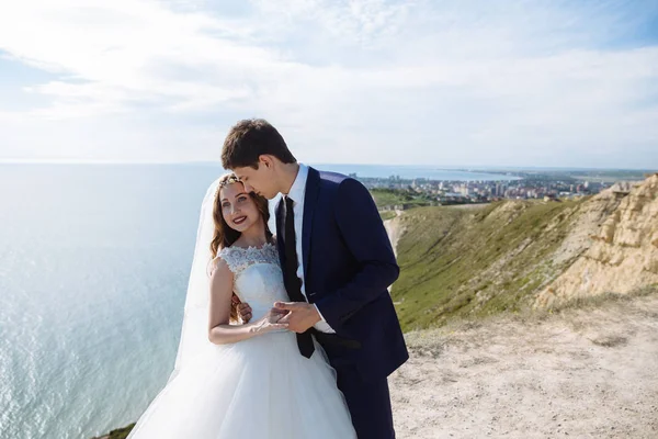 Elegante novio sostiene novia de la mano, abraza y quiere besar. Recién casados el día de la boda en la montaña con vista al mar —  Fotos de Stock