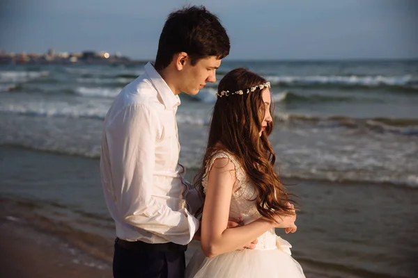 Vőlegény ölelést gyönyörű menyasszony a sugarak naplemente ocean beach. Ifjú házasok időt együtt az esküvő napján — Stock Fotó