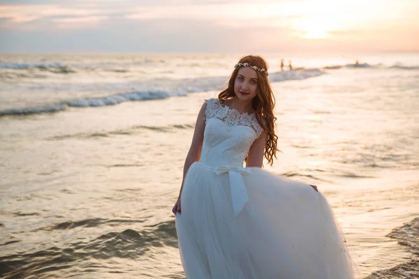 Boldog menyasszony elegáns esküvői ruhát és szép frizurát a ocean beach. Menyasszony séta a tengerparton naplementekor — Stock Fotó
