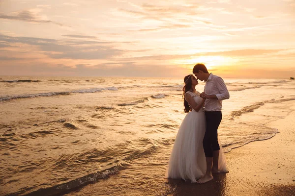 Sposa e sposo che si abbracciano e si tengono per mano in uno splendido sfondo al tramonto. Sposi al giorno del matrimonio sulla spiaggia dell'oceano — Foto Stock