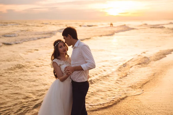 Buon sposo e sposa che si abbracciano sulla spiaggia dell'oceano al tramonto. Coppia romantica sposi il giorno del loro matrimonio . — Foto Stock