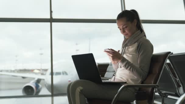 Vista lateral da menina bonita usa telefone e laptop para trabalhar no aeroporto enquanto espera embarque na sala de embarque — Vídeo de Stock