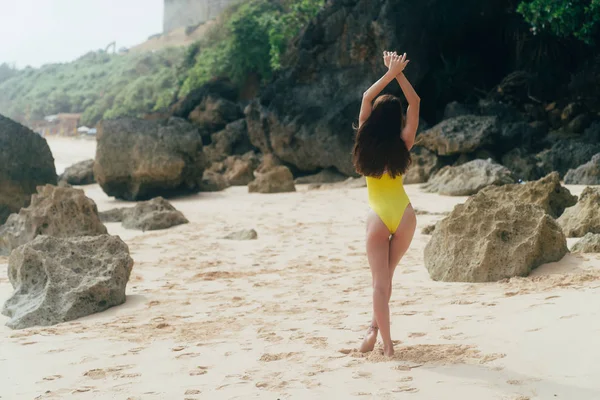 Vista trasera delgada chica bronceada descansando en la playa de arena con rocas en el fondo —  Fotos de Stock