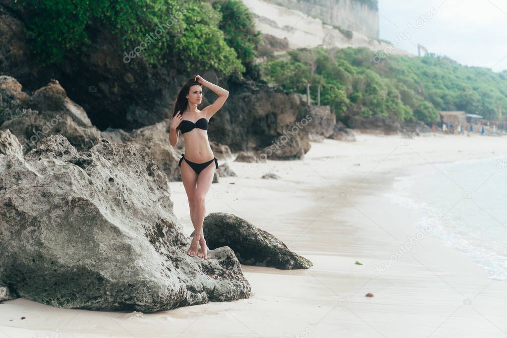 Sexy tanned brunette girl in black swimwear sunbathes on beach with rocks.