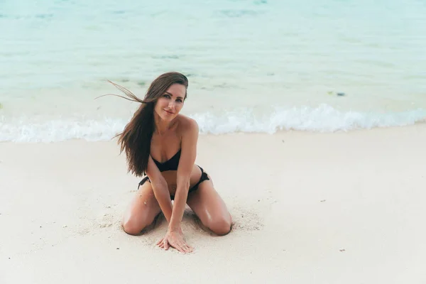 Sexy chica bronceada en traje de baño negro posando en la playa de arena cerca del océano. Hermosa modelo broncea y descansa —  Fotos de Stock