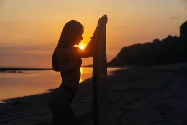 Silueta de chica delgada con tabla de surf en las manos en la playa en el fondo de la hermosa puesta de sol — Foto de Stock